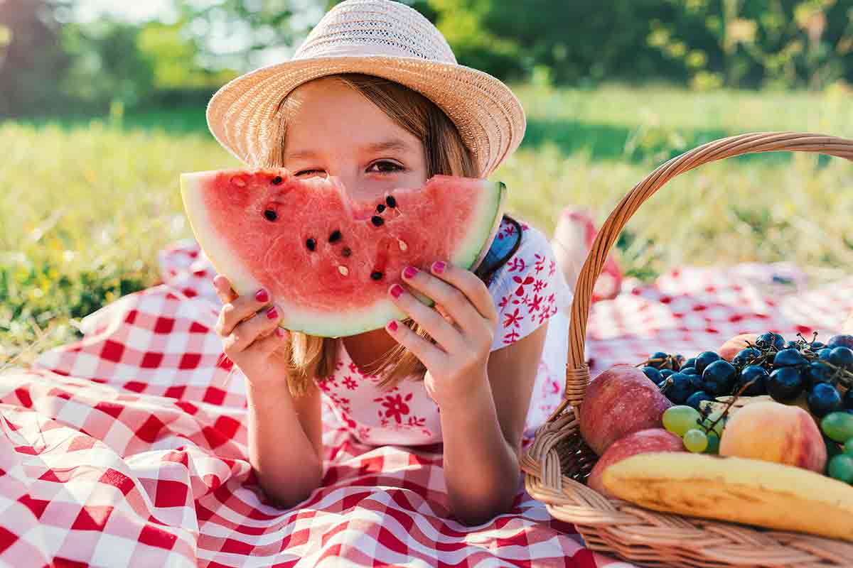 Girl on picnic blanket looking from behind a slice of watermelon. 
