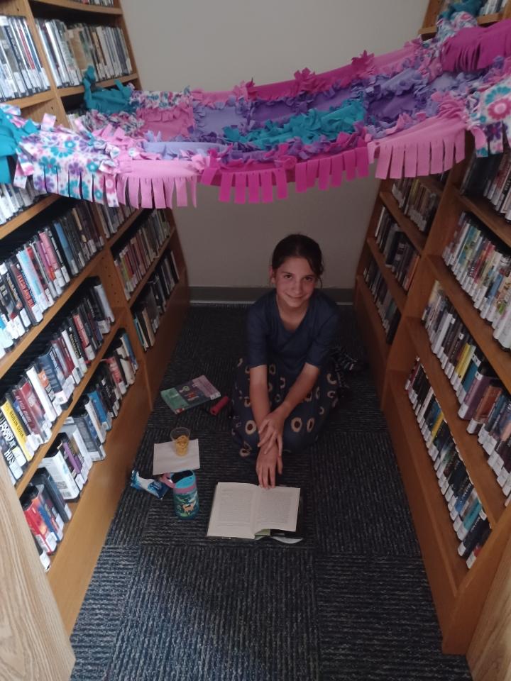 Little girl sitting on floor under a blanket layed across bookshelves above