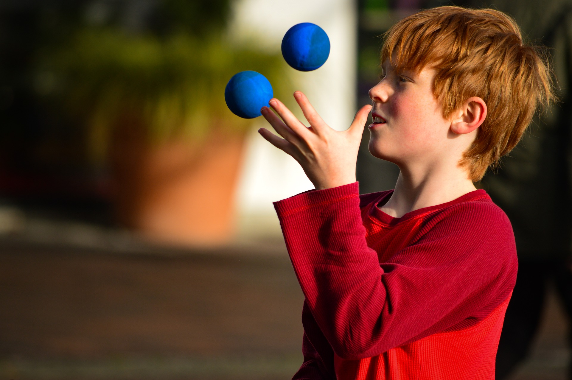 ginger-haired boy wearing a red shirt juggling two blue balls