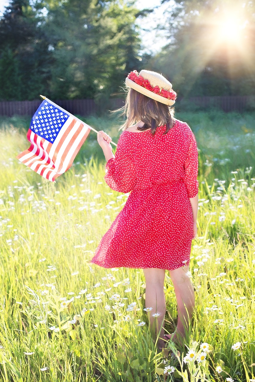 girl wearing a red dress standing in a field of tall grass holding an American flag