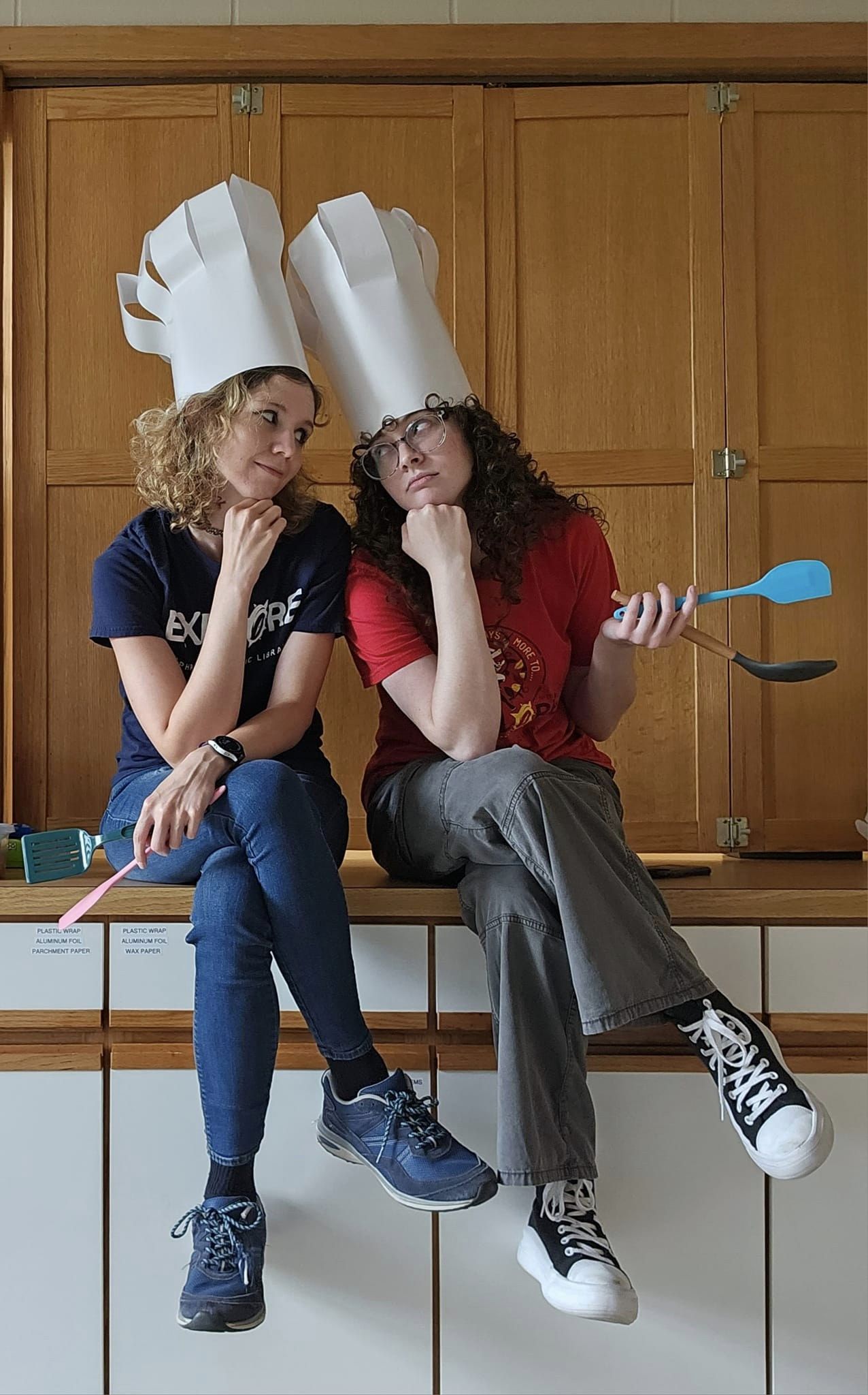 Laura and Ascher wearing chef hats and looking both tired and amused while holding spatulas