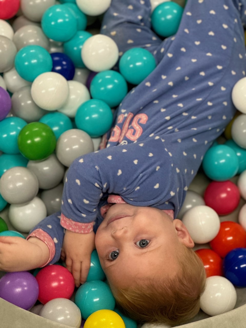 Photo of child wearing blue outfit with white dots. They are laying in a ball pit of colorful balls. 