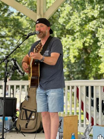 Photo of man in gray shirt and blue jean shorts holding a guitar