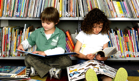 children sitting on library floor, arm to arm, both reading a book