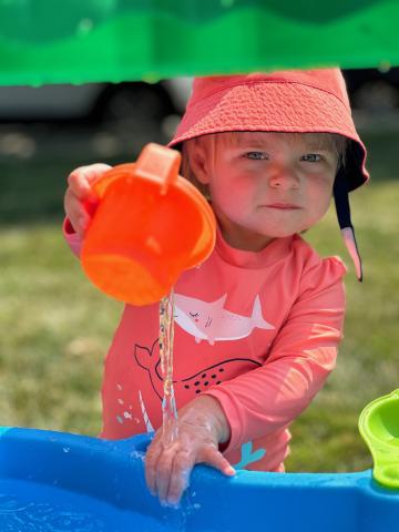 Child in coral colored swim wear holding an orange cup standing at a blue water table with green tree top. 