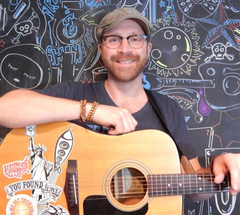 Mark DeRose sitting in front of a colorfully decorated blackboard with his guitar