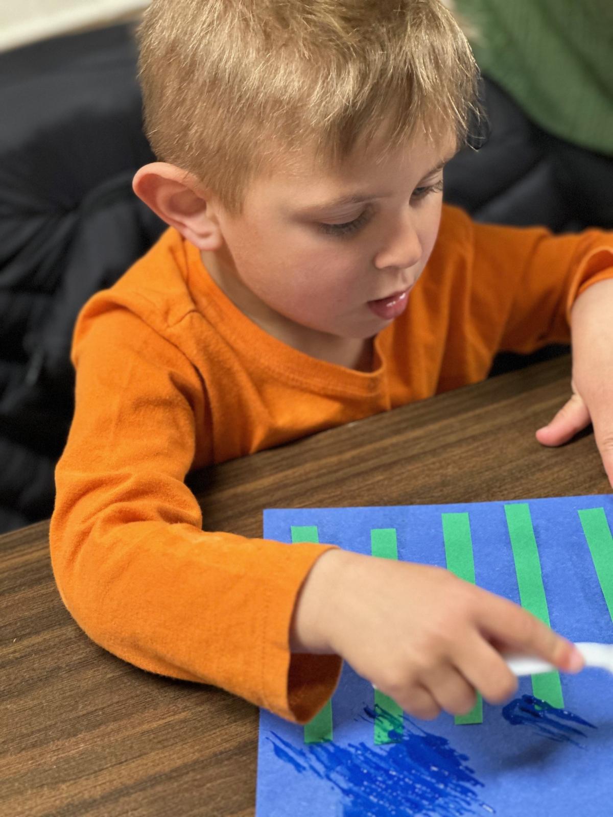 Child wearing orange shirt painting on a blue paper during story time. 