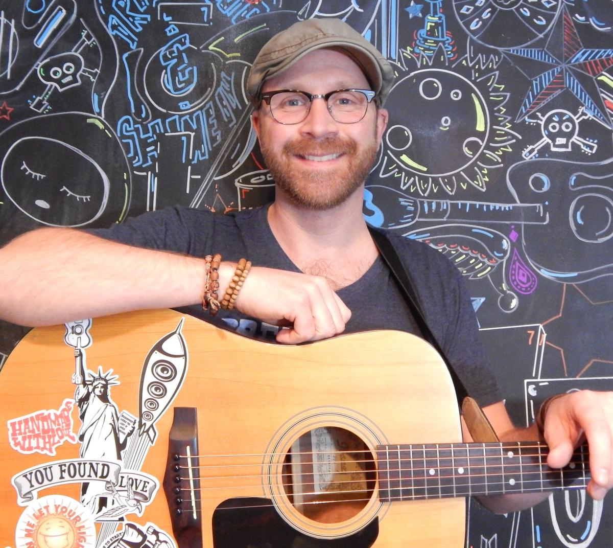 Mark DeRose sitting in front of a colorfully decorated blackboard with his guitar